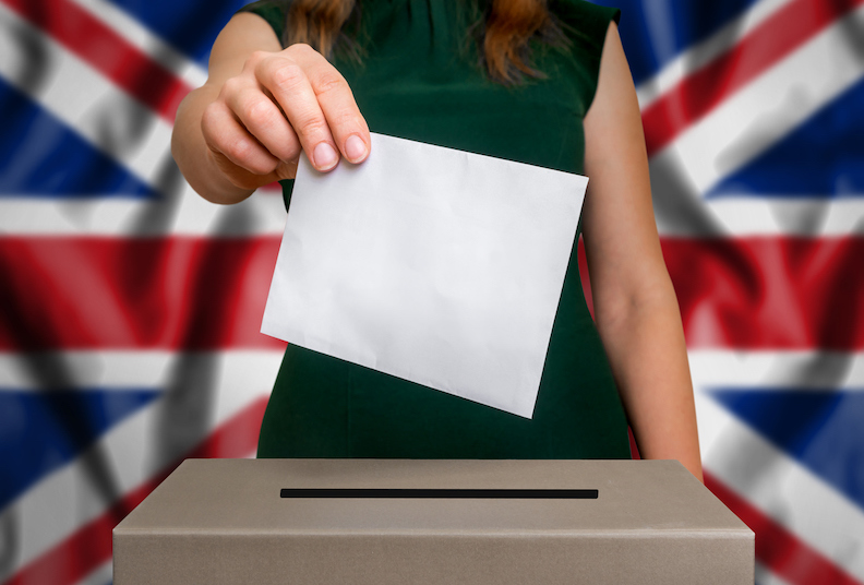 Election in United Kingdom - voting at the ballot box. The hand of woman putting her vote in the ballot box. Flag of United Kingdom on background.